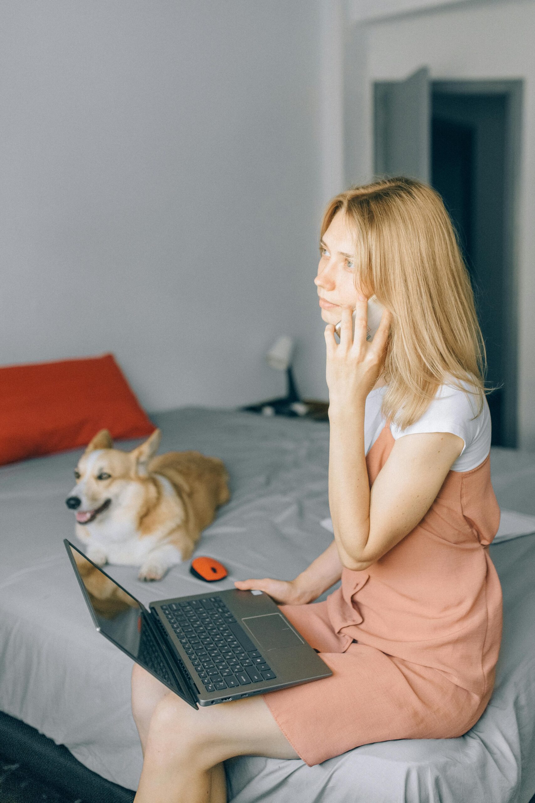 Young woman multitasking on laptop and phone with cute corgi on the bed.